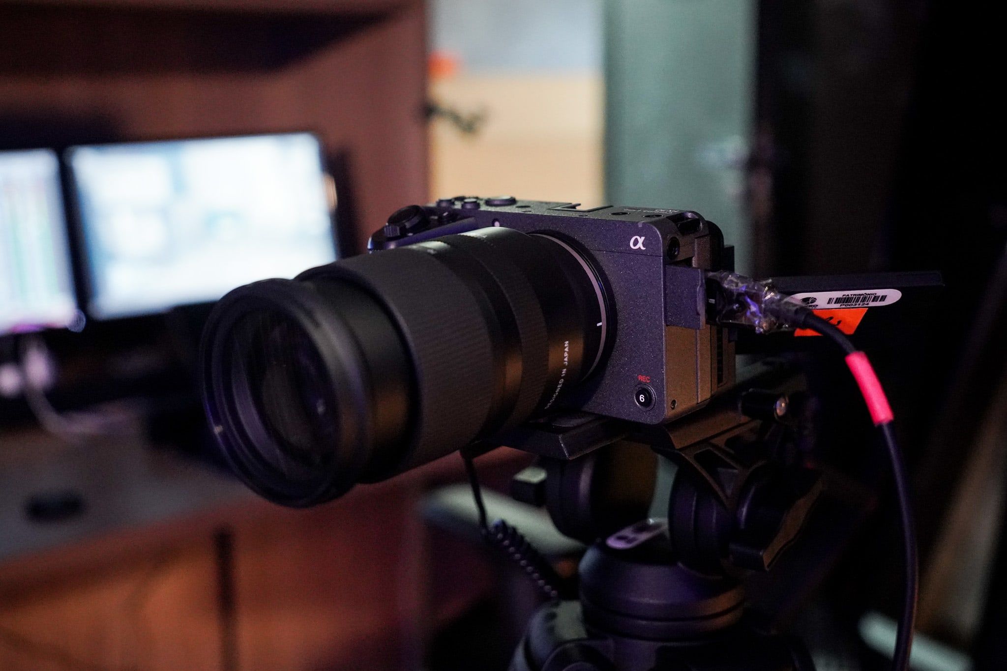 Câmera Sony com lente zoom no tripé, com dois monitores desfocados ao fundo.
