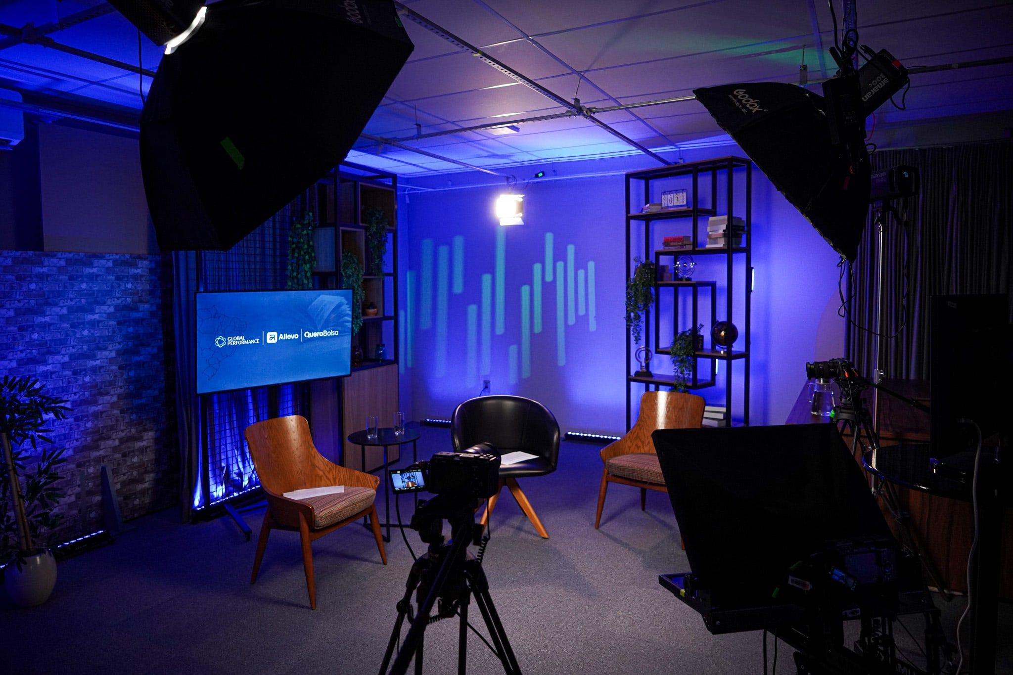 Set do estúdio sob luz azul e roxa: três poltronas, uma TV na parede, a câmera no tripé em primeiro plano, o teleprompter à direita e duas softboxes no alto.